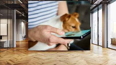 Woman using mobile phone at home. Female sitting on sofa with dog and browsing social networks on smartphone Wall mural