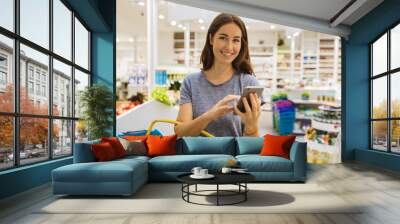 Casual young female, using her cellphone in modern supermarket. Looking at camera Wall mural