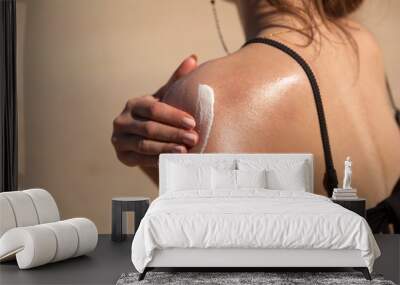 A young woman applying sun cream or sunscreen on her tanned shoulder to protect her skin from the sun. Shot on a sunny day with blurry sand in the background. Wall mural