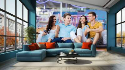 A company of good-looking friends is laughing and sitting on the bench with cushions in the nice summer cafe. Entertainment, having good time. Friendship. Wall mural