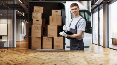 A young handsome smiling worker wearing uniform is standing next to the van full of boxes holding a clipboard in his hands. House move, mover service. Wall mural