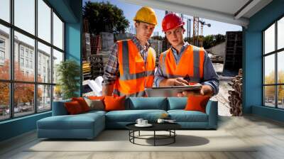 Architect  and structural engineer dressed in orange work vests and  helmets discuss a building project on the tablet on the open air building site with construction material Wall mural