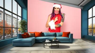 Girl with purple hair tips and tattoo on her arm dressed in red swimsuit, Santa's hat and white fur bracelets holding a lollipop in front of her face on the background of pink wall Wall mural