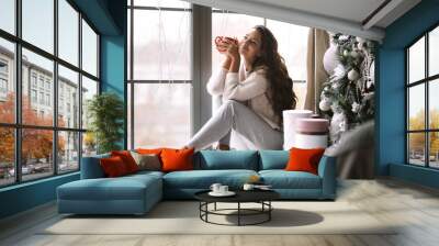 Nice dark-haired girl dressed in pants, sweater and warm slippers holds a red cup sitting on the windowsill of a panoramic window in the room next to the New Year tree and gifts Wall mural