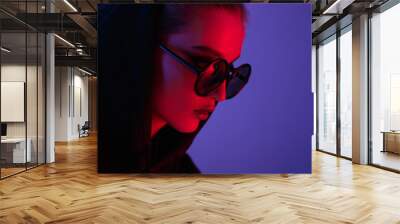 Portrait of fashion young girl in a black sweater with a hood and round sunglasses in red and blue neon light in the studio Wall mural