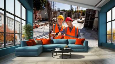 Structural engineer and architect dressed in orange work vests and  hard bats  discuss the construction process on the open building site with construction material Wall mural