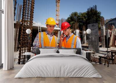 Structural engineer and architect dressed in orange work vests and  hard hats discuss the construction process by the phone and use tablet  on the open building site Wall mural