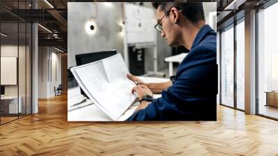 Stylish dark-haired architect in glasses and in a blue jacket is working with documents on the desk in the office Wall mural