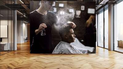 The barber dressed in a black clothes is doing the hair styling to the stylish black-haired  man sitting in the armchair in a barbershop Wall mural