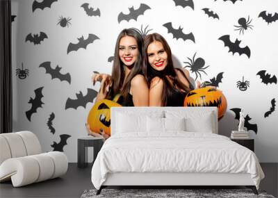 Two beautiful brunette women in black dresses have fun with jack-o-lanterns on a white background with bats and spiders Wall mural