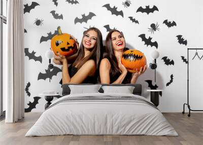 Two charming brunette women in black dresses with red lipstick hold Halloween pumpkins on a white background with bats and spiders Wall mural