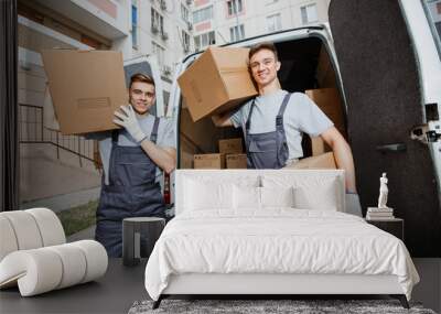 Two young handsome smiling workers wearing uniforms are standing in front of the van full of boxes holding boxes in their hands. The block of flats is in the background. House move, mover service. Wall mural