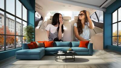 Two young pretty slim girls with long hair,dressed in casual style, are sitting in the back seat of a fancy and chatting. Wall mural