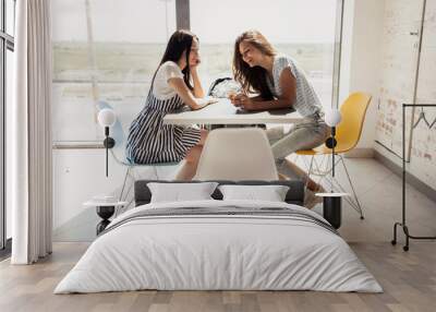 Two youthful pretty slim girls with dark hair,wearing casual outfit, sit at the table next to each other and chat in a modern office. Wall mural