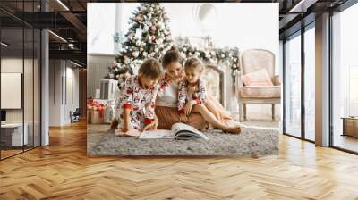 Young beautiful mother with two little daughters sit on the carpet and read book near the New Year's tree with gifts in the light cozy room with armchair and fireplace Wall mural