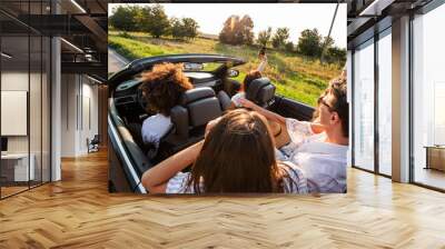 Young girls and guys are sitting in a black cabriolet and making selfie on a warm sunny day. Wall mural