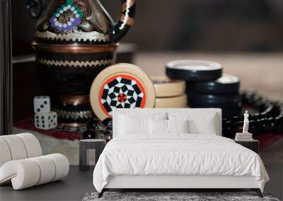 A beautiful teapot stands on a rug next to stacks of black and white patterned backgammon chips, dice with numbers and rosaries: close up Wall mural