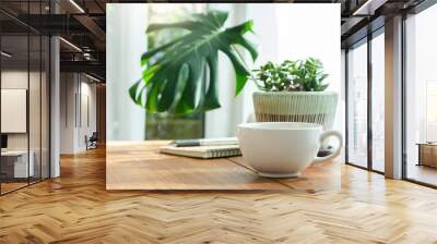 Soft focus of white coffee cup with small trees and green leaf in vase on wooden table near bright window Wall mural