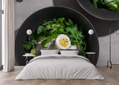 Green summer salad with arugula, cucumbers and egg on black plate. Top view food with Sunny light. Wall mural