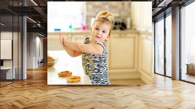 Little toddler kid having lunch in the warm sunny kitchen. Blonde girl with funny ponytail playing with two tasty bagels Wall mural