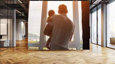 Father holding toddler daughter in arms opens curtains of panoramic window together. Parent and child enjoy morning view of city from home backside view Wall mural