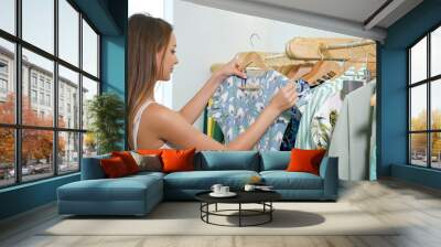 slim woman with loose fair hair holds wooden hanger and looks at blue summer dress near rack in shop close view Wall mural