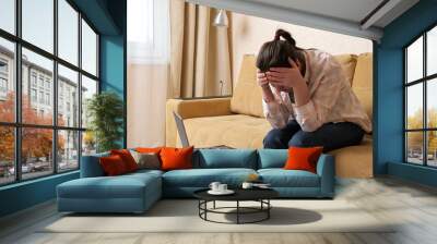 Upset woman tries to calculate high payments on modern mobile phone and holds head in frustration sitting on beige sofa in light living room Wall mural