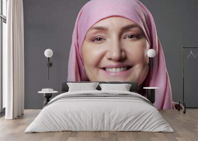 Young Muslim woman in light pink-colored hijab smiles widely posing for camera with confidence in studio room at professional illumination closeup. Wall mural