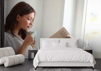 young woman at home sitting near window relaxing in her living room reading book and drinking coffee Wall mural