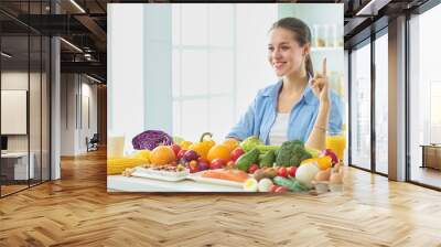 happy young housewife sitting in the kitchen preparing food from a pile of diverse fresh organic fruits and vegetables, selective focus Wall mural