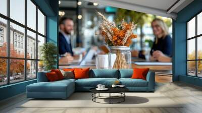 A close-up of a glass jar vase filled with dried flowers and wheat stands on a wooden meeting table, with blurred professionals discussing in the background. Wall mural