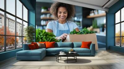 A happy woman in casual attire packages fresh greens in a modern kitchen setting, holding a cardboard box filled with leafy vegetables, surrounded by a lively kitchen atmosphere. Wall mural