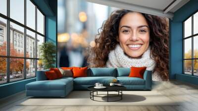 A happy woman with curly hair and a warm scarf, smiling as she enjoys a winter day in the city, reflecting warmth and positivity amidst the chilly urban environment. Wall mural