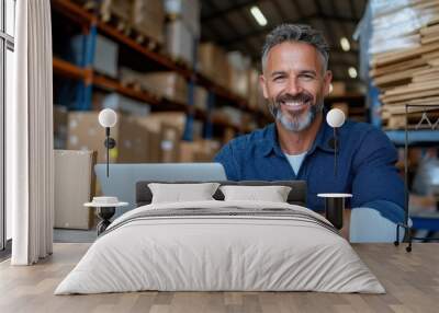 A man with gray hair and beard smiling while working on a laptop computer inside a warehouse filled with shelves and boxes, projecting a professional attitude. Wall mural