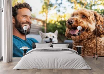 A smiling man enjoys outdoor time with two happy dogs, capturing the essence of joy, companionship, and warmth in a brightly lit natural setting filled with greenery and sunlight. Wall mural