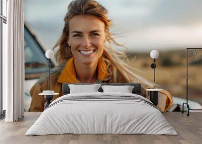 A smiling woman with flowing hair and dressed in casual country attire leans on a car in an open countryside setting with golden, grassy fields in the background. Wall mural