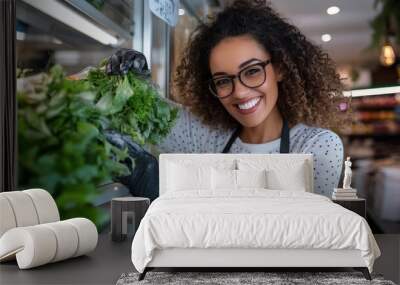 A woman with glasses and curly hair, smiling as she organizes fresh greens in a grocery store, showcasing freshness and attentive service in a retail environment. Wall mural