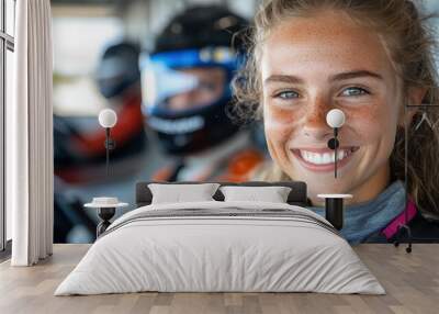 A young female racer with freckles beams confidently at the camera, seated in her go-kart with a helmet, amid a background of bustling karting activity. Wall mural