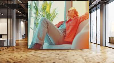 Relaxed man in casual attire leaning back in a sunlit room enjoying a moment of tranquility with lush green plants and warm natural light streaming through large windows Wall mural