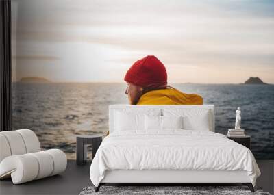 Alone man traveling on ship and looking at sunset sea and foggy mountain on skyline. Hipster traveler wearing yellow raincoat and red hat enjoying beautiful ocean after storm Wall mural