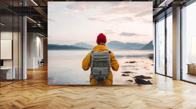 Back view of male tourist with rucksack standing on coast in front of great mountain massif while journey.  Man traveler wearing yellow jacket with backpack explore nature. Wanderlust lifestyle Wall mural