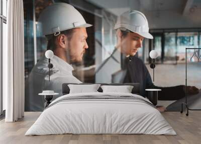 Building worker and architect discussing build drawing on construction site. Two industrial engineers wearing safety hard hat have meeting on commercial building structure Wall mural