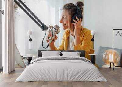 Cheerful woman podcaster recording her voice into microphone. Female radio host streaming podcast using microphone and laptop at his home studio Wall mural