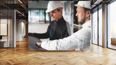 Industrial architect discussing build drawing with worker wearing safety hard hat. Two building engineers lookin at blueprint on construction site Wall mural