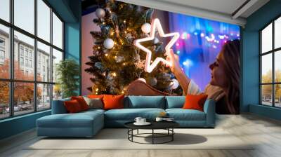 Cheerful young woman sitting next to christmas tree and holding glowing LED star in dark cozy room at xmas eve Wall mural