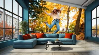 Happy young woman jumping with raised arms on colorful autumn leaves city background Wall mural