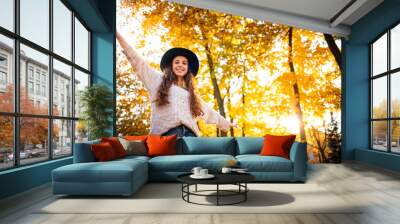 Joyful stylish young woman with raised arms walking on colorful autumn city street at sunny day Wall mural