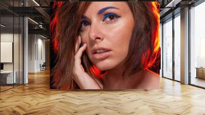 Unretouched portrait of a young pretty caucasian brunette girl posing in a studio on black background with bright orange backlight. She has messy hair and bright blue eye liner and beige lip gloss. Wall mural