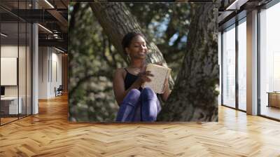 Sitting high up on a tree branch, a person writes a diary entry in their journal. They are African American, wearing casual athletic wear, and holding a pen and notebook. Wall mural