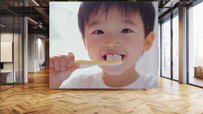 A young boy smiles while brushing his teeth with a yellow toothbrush. Wall mural